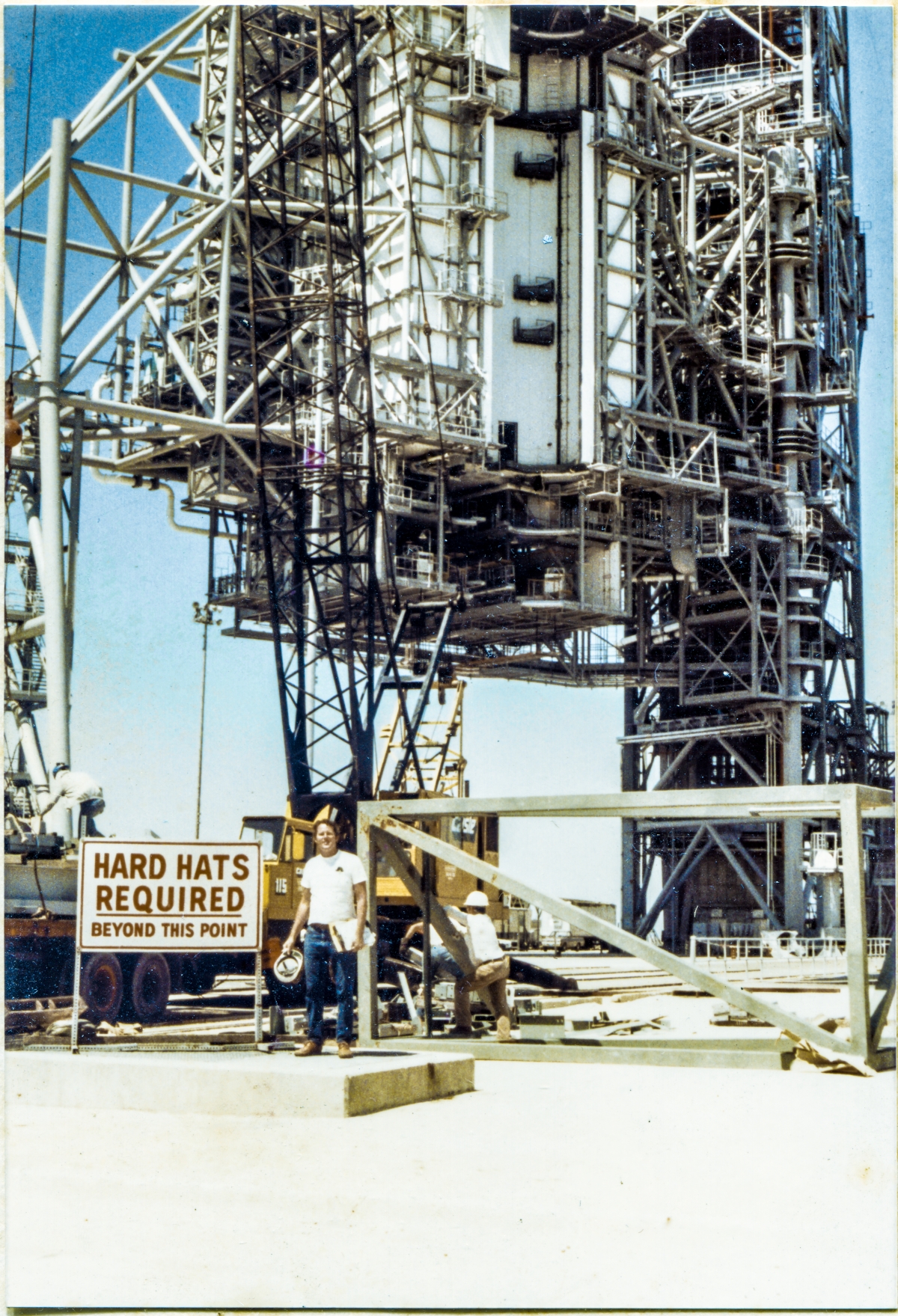 Image 055. Our earliest look at James MacLaren at Space Shuttle Launch Complex 39-B, Kennedy Space Center, Florida, after returning to the jobsite following his layoff from Sheffield Steel, and his subsequent hiring by Ivey Steel. In the far distance, the RSS and FSS loom into the sky, overflowing the frame, sensibly-complete in general appearance, but far from complete for the purposes which they were designed and constructed for, servicing the Space Shuttle, prior to launch. In the near distance, the yellow 90-ton P&H crane is being used to offload more steel from the back of a flatbed semitrailer. Union Ironworkers from Local 808 work the load from on top of the trailer and on the ground next to it, while the white-shirted truck driver lounges against the lopped-off tube steel top of the East Stair Tower. In his left hand, MacLaren grips a clipboard holding the sheets of the shipping list that came with the load of iron, with a yellow notepad behind it. In his right hand, he holds his hard hat, and if you look very close, you can also see a yellow highlighter in that hand for marking up the shipping list indicating items which have been checked and verified as being included in this delivery. On his right hip, a 25-foot tape measure that stayed with him wherever he went on the Pad Deck or on the Towers. Another day on the job. Photo by James MacLaren.